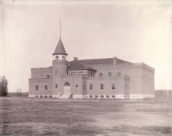 A historic, rectangular brick building with a castle-like appearance, featuring a central tower with a conical roof. The building stands on a bare, grassy field under an overcast sky.