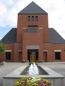 A brick church with a triangular roof and a central entrance is shown. In the foreground, a rectangular fountain with multiple water jets is surrounded by colorful flowers. The sky is partly cloudy.