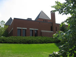 A modern brick building with slanted roofs surrounded by greenery. The structure features large windows, a chimney, and is set against a clear sky. Bushes and a well-maintained lawn are in the foreground.