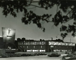 A black-and-white image of a building at night, with illuminated windows and a sign displaying a logo. The foreground contains parked cars, and tree branches frame the top of the image.