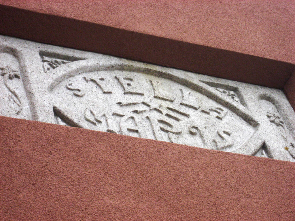 Close-up of a carved stone embedded in a red wall. The stone features ornate carvings with the word "STELLE" prominently displayed in the center, surrounded by decorative patterns.