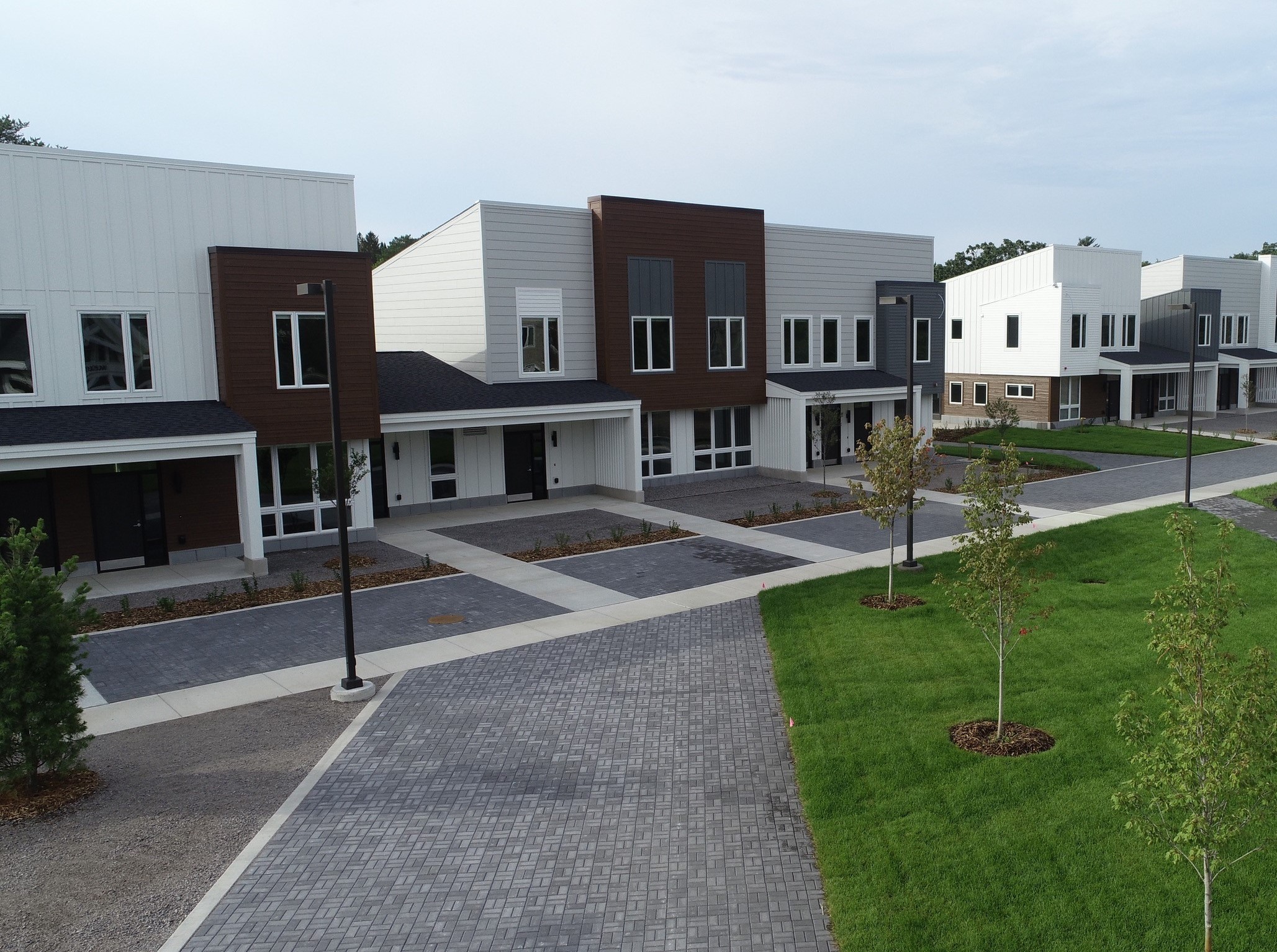 A modern residential street with newly constructed houses featuring white and brown facades. The homes have flat roofs and large windows. The street is paved with bricks and landscaped with small trees and green grass patches. The sky is clear.