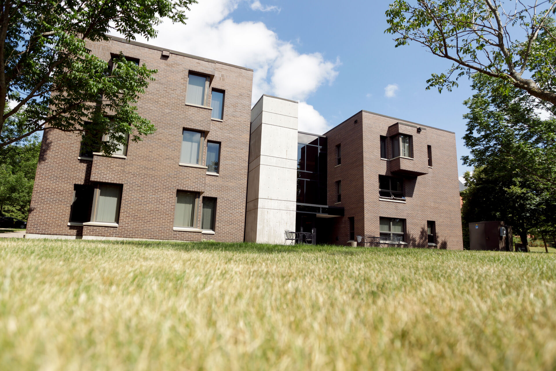 A modern brick apartment building with large windows sits on a grassy lawn. The structure has two main sections connected by a central white facade. Green trees frame the scene under a partly cloudy blue sky.