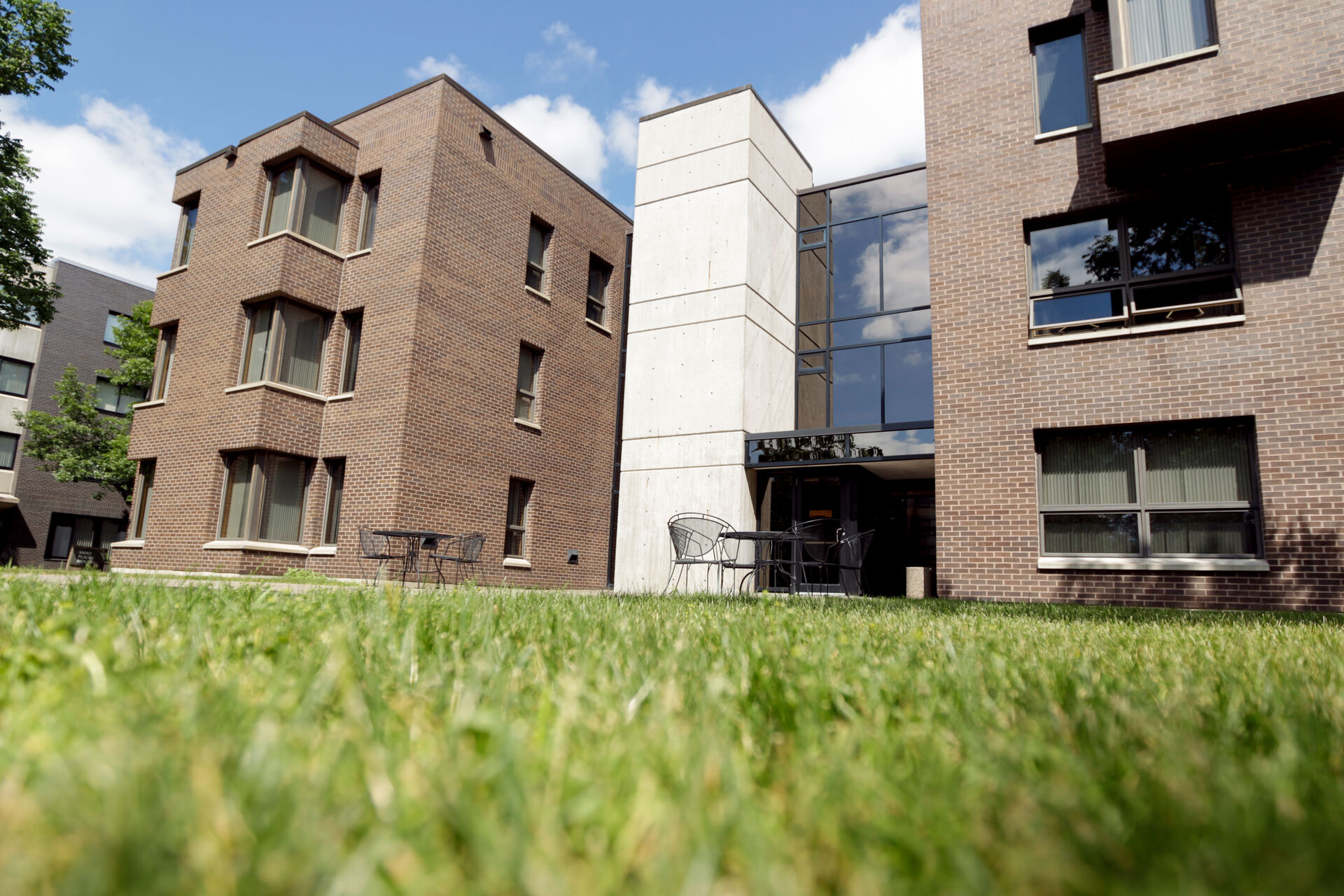 A low-angle view of a modern brick building with large windows and a central white concrete section. The foreground features a well-maintained lawn, while the sky is blue with a few clouds. Black patio chairs and tables are visible near the entrance.