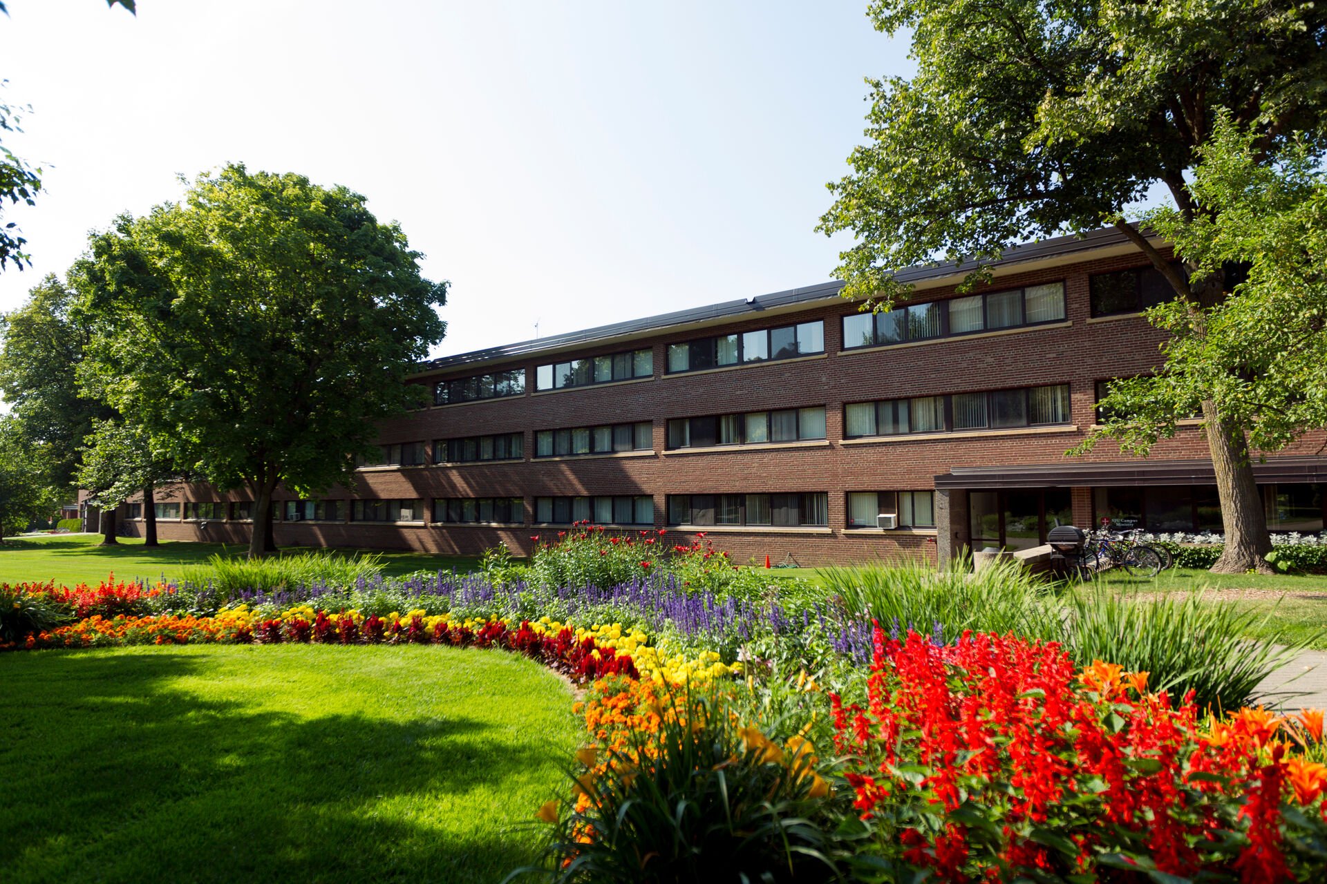 A three-story brick building with large windows is surrounded by lush green trees and a vibrant garden filled with colorful flowers, including red, yellow, and purple blossoms. The sky is clear and sunny.