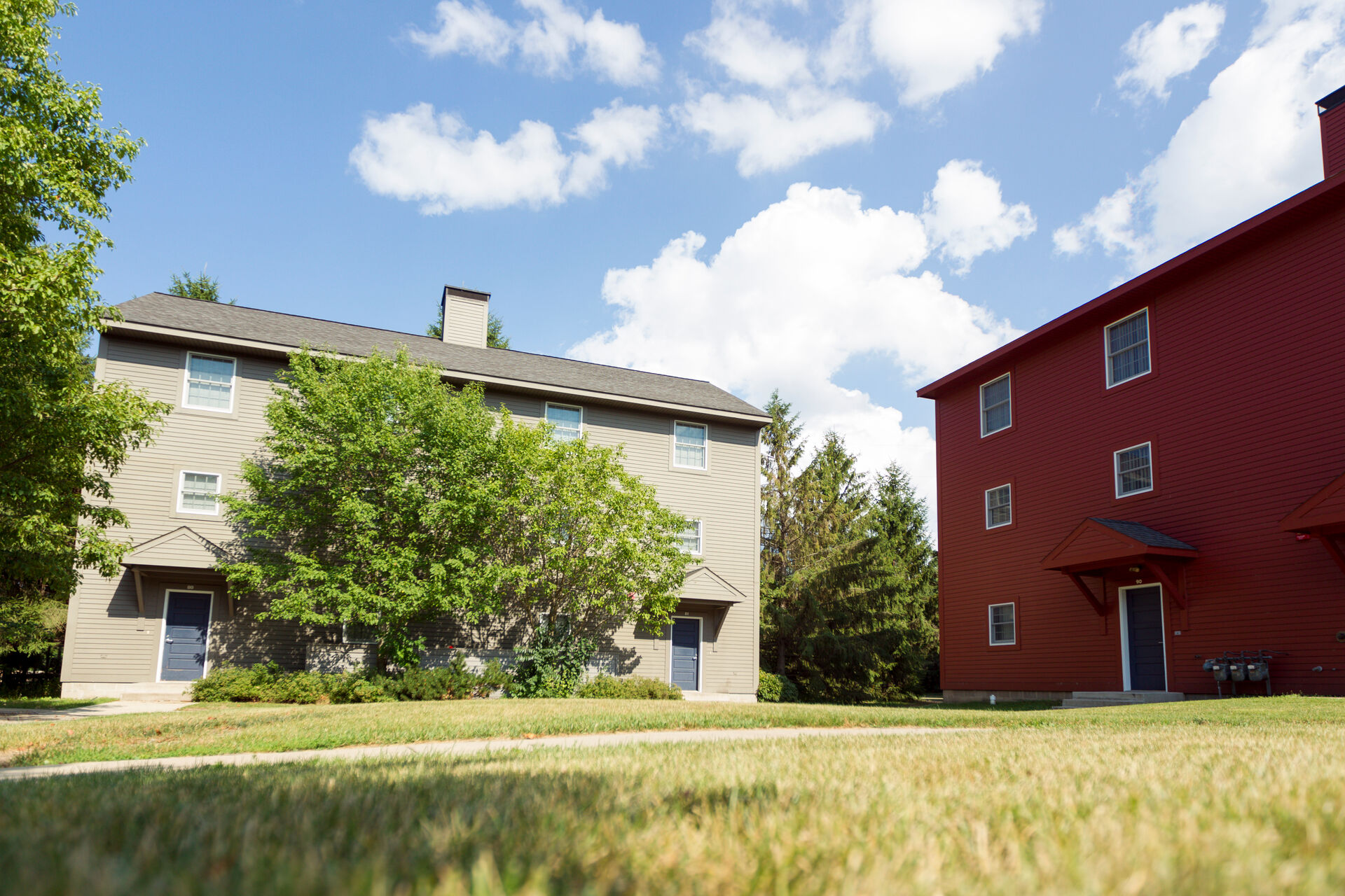 Two residential buildings with sloped roofs under a blue sky. The building on the left is beige with a small tree in front, and the one on the right is red. Both are surrounded by trimmed grass and a few scattered trees.