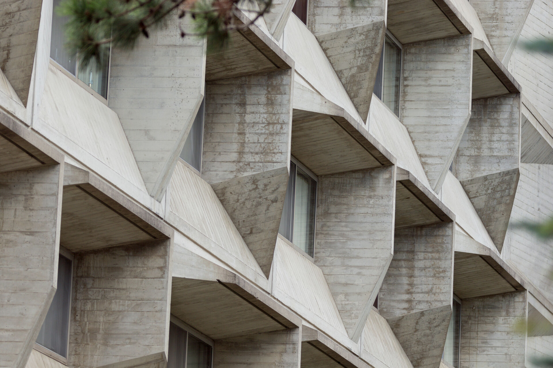 Close-up of a modern building facade featuring geometric, angular concrete structures. The design creates a pattern of protruding triangular shapes with windows in between. The photo is taken at an angle, highlighting the building's texture and symmetry.