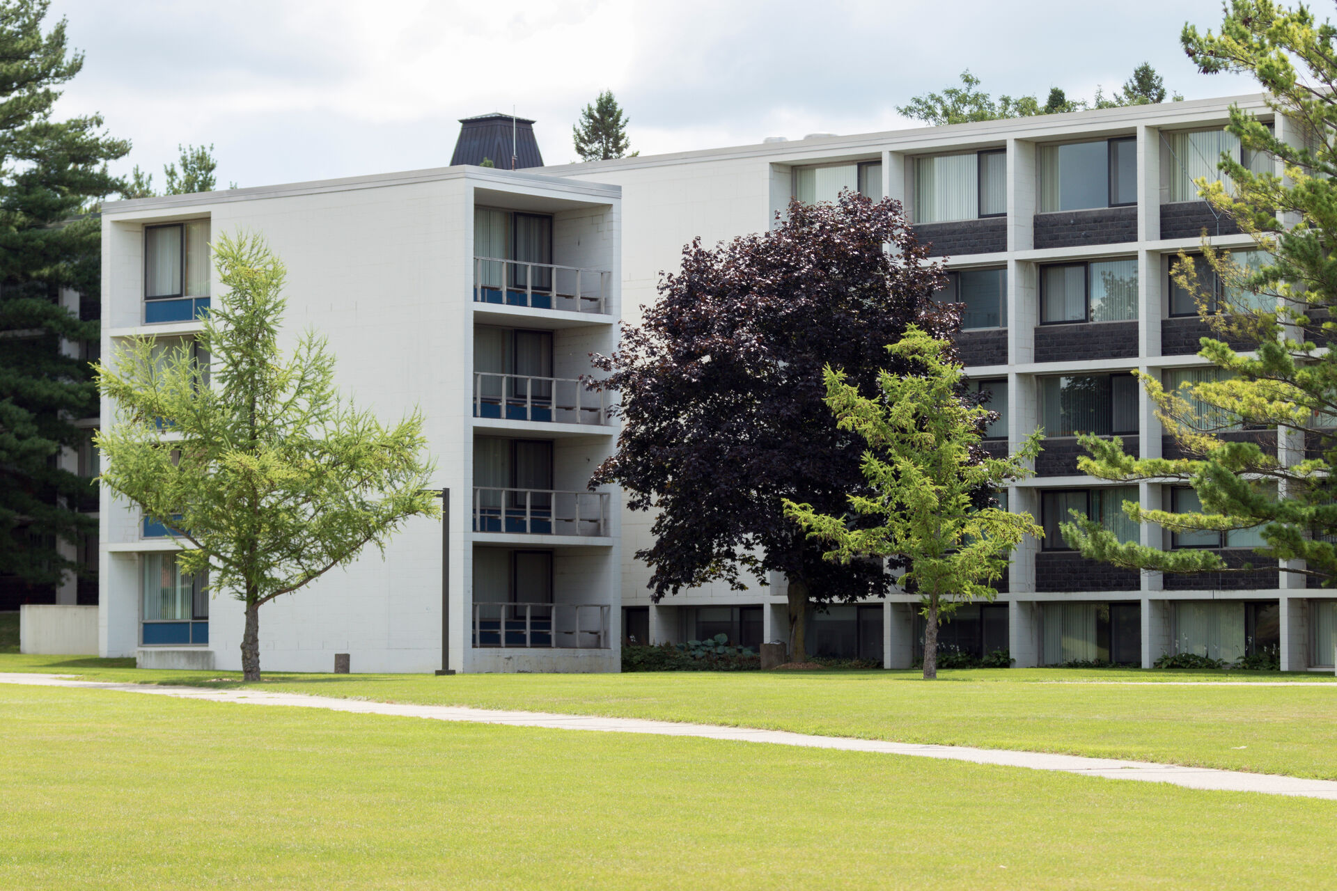 A modern four-story apartment building with balconies, surrounded by well-maintained grass and trees. The structure features a minimalist design with large windows and a flat roof. The sky is partly cloudy.