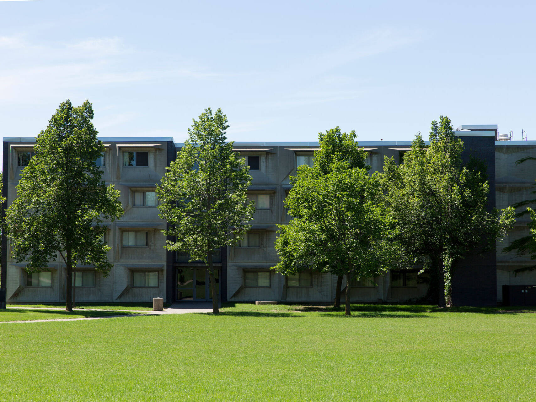 A four-story concrete residential building surrounded by lush green grass and trees under a clear blue sky. The structure features rows of windows and has a symmetrical design.