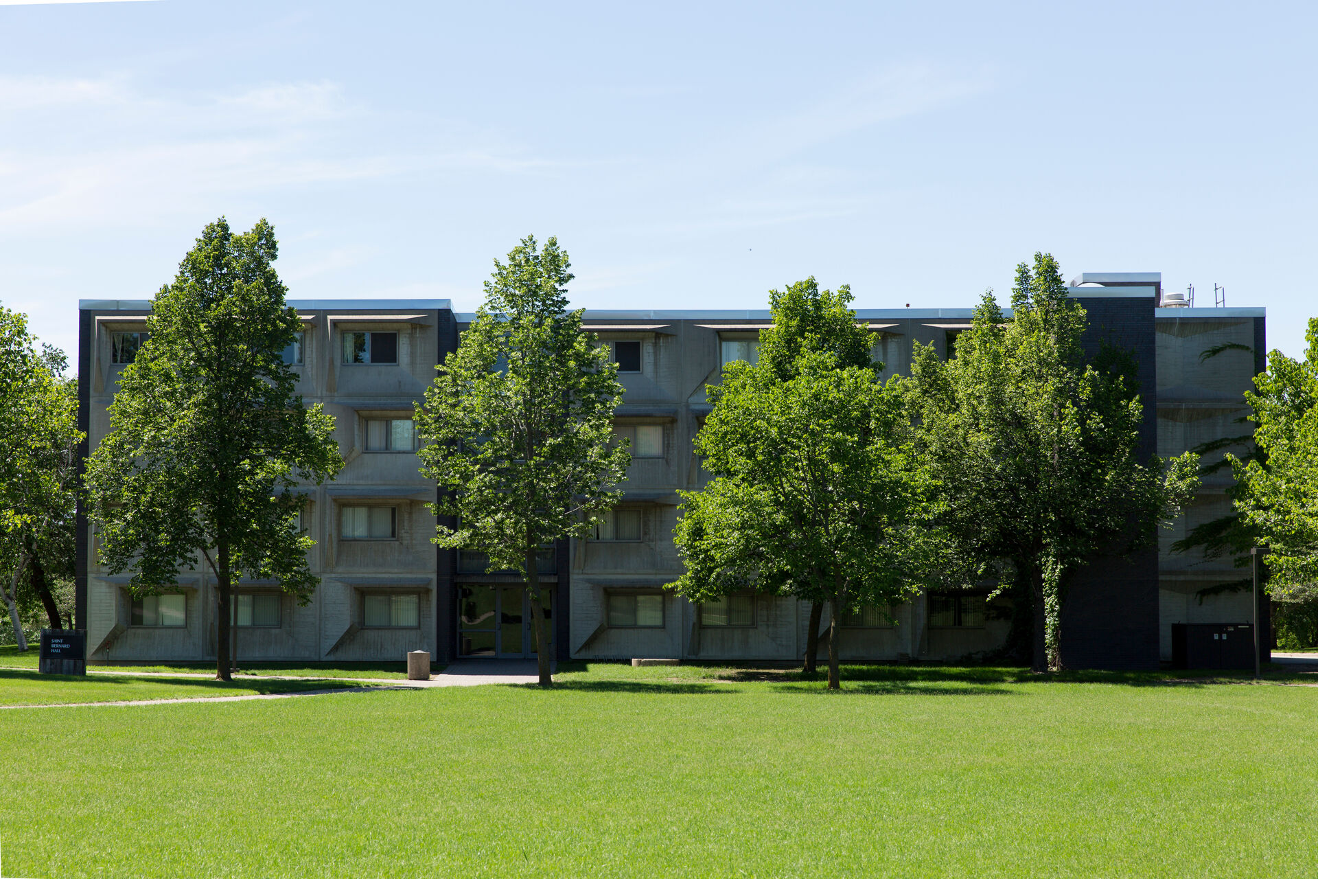 A four-story concrete residential building surrounded by lush green grass and trees under a clear blue sky. The structure features rows of windows and has a symmetrical design.