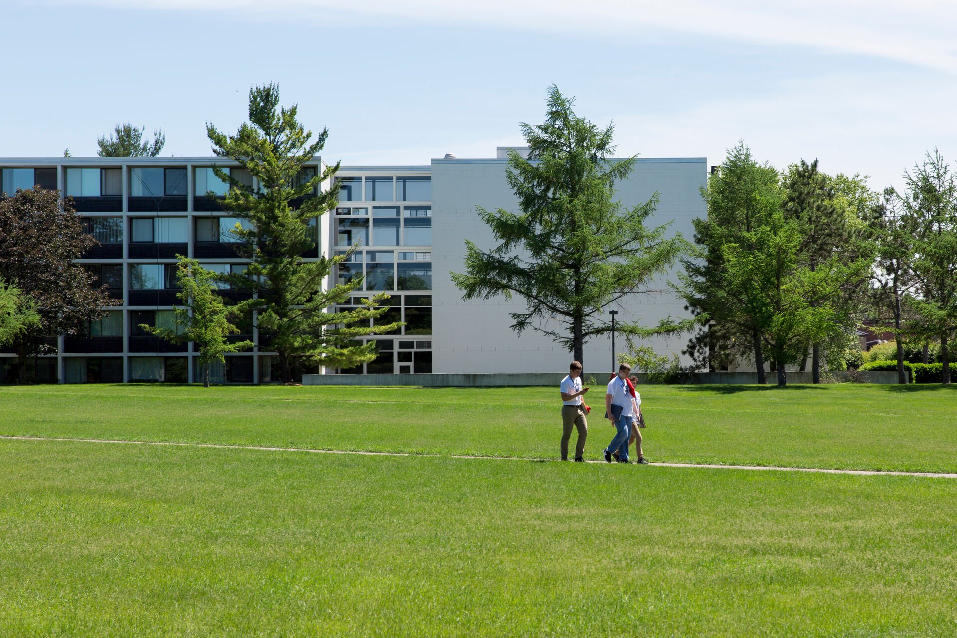 Two people walk along a path through a grassy field with a modern building in the background. The building has large windows and is surrounded by trees. The sky is clear and sunny.