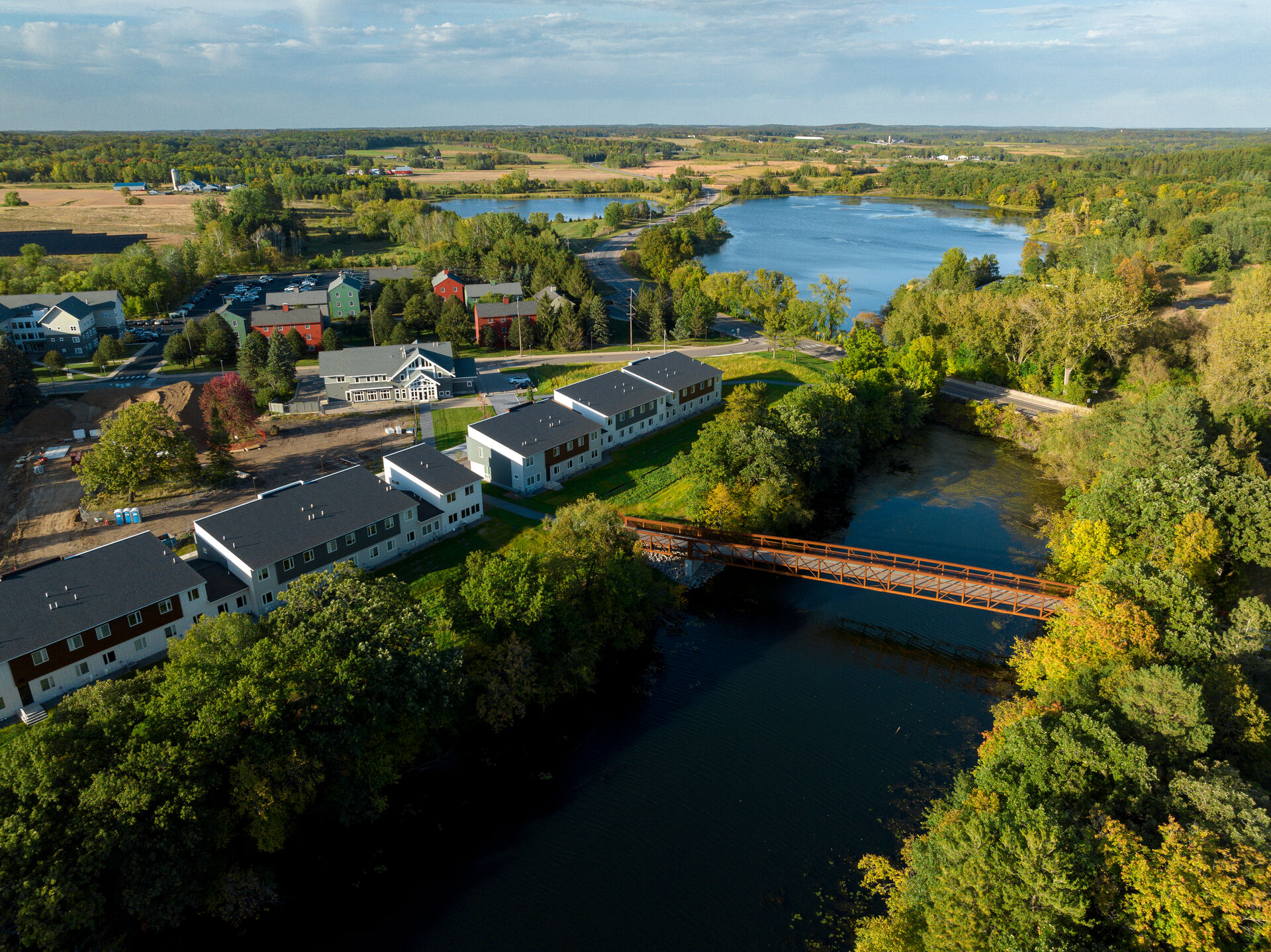 Aerial view of a riverside community with multiple residential buildings surrounded by trees. A bridge crosses the river, and fields and forests are visible in the background under a partly cloudy sky.