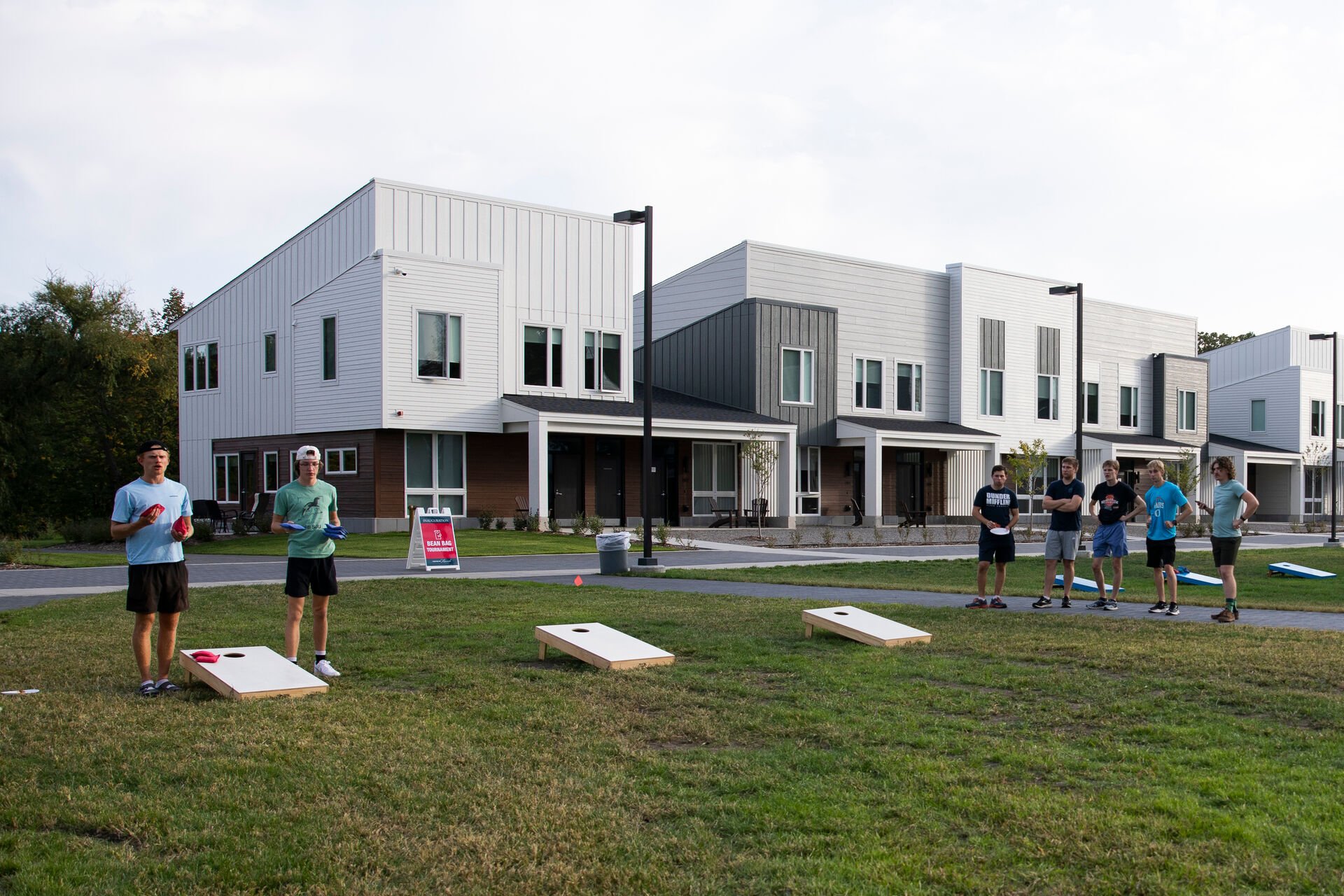 A group of people playing a game of cornhole on a grassy area in front of modern townhouses. Four wooden boards are set up, and several individuals are watching or participating in the game on a sunny day.