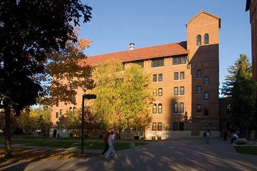 A large red-brick building with a tall tower, surrounded by trees with autumn foliage. There is a pathway in front where a few people are walking. The scene is bathed in the warm light of the setting sun.