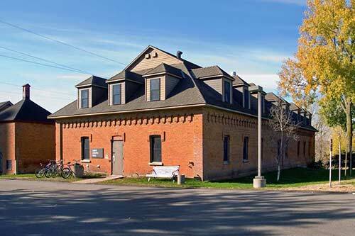 A rectangular, two-story brick building with dormer windows and a gabled roof. Bicycles are parked near the entrance, and there is a white bench beside the door. It's set in a sunny landscape with trees, including one with yellow autumn leaves.