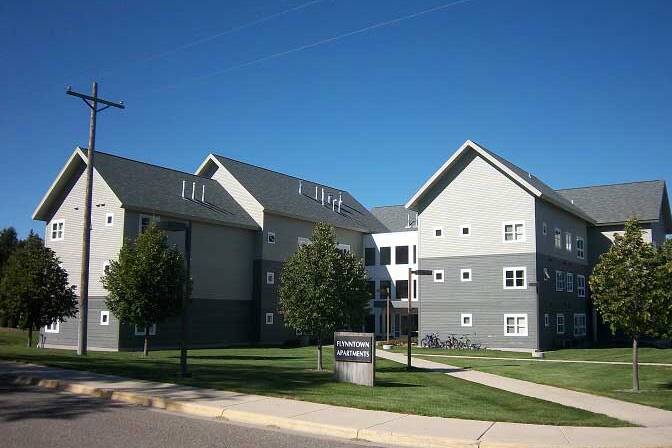 A modern apartment complex with gray siding and white trim under a clear blue sky. The buildings have multiple stories with several small windows. Lush green lawns and trees surround the structures. A sidewalk curves in the foreground.