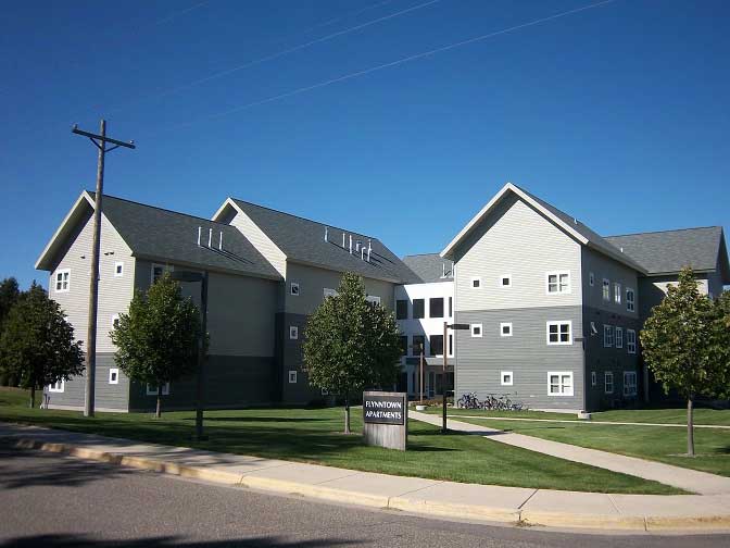 A modern apartment complex with gray siding and white trim under a clear blue sky. The buildings have multiple stories with several small windows. Lush green lawns and trees surround the structures. A sidewalk curves in the foreground.