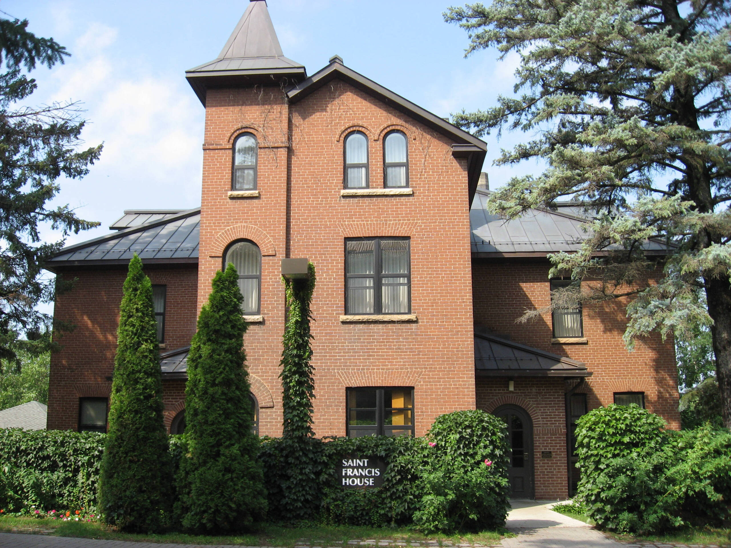 A brick building with arched windows and a turret, partially covered by tall green shrubs. A sign reads "Saint Francis House" in front of the building. Trees surround the structure under a blue sky with clouds.