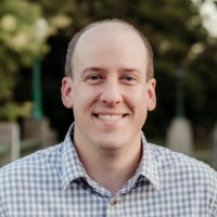 A person with a shaved head and a checked shirt smiles against an outdoor background with blurred greenery.