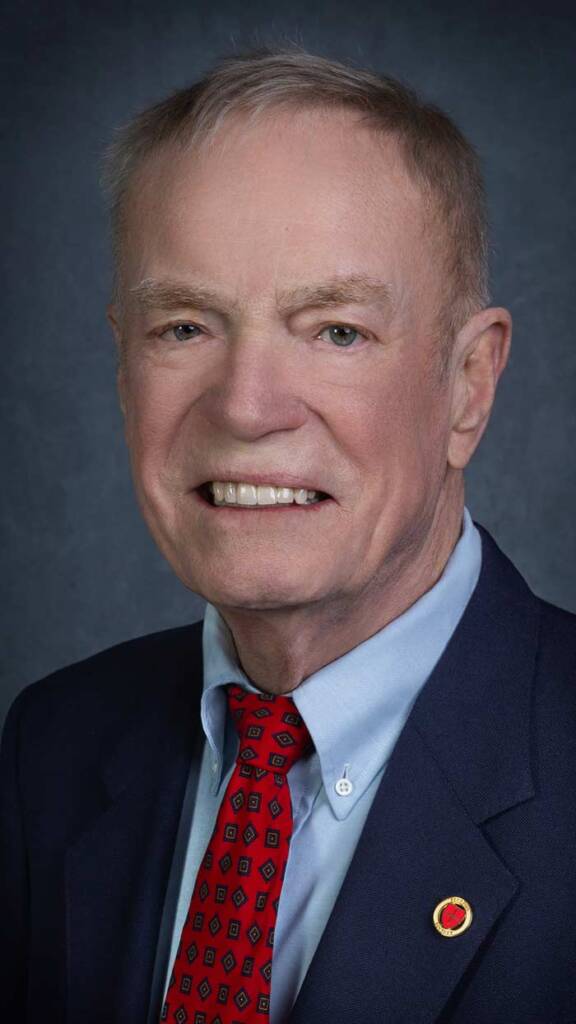 An older man wearing a navy suit, light blue shirt, and red patterned tie. He has short gray hair and is smiling. The background is a plain dark color.