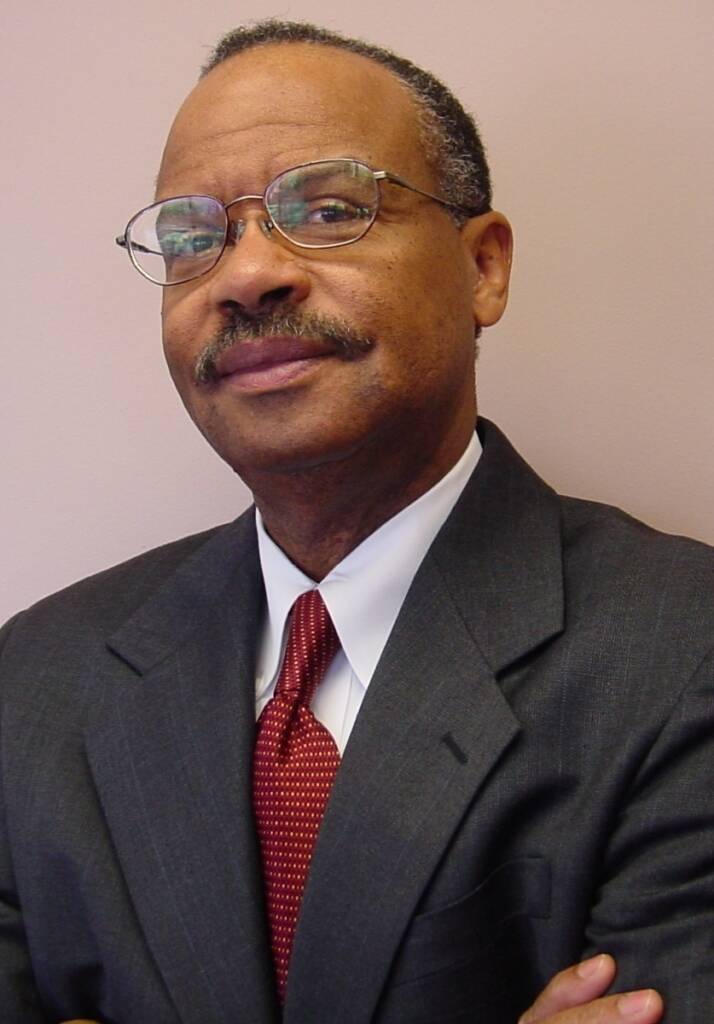 A person in a suit and tie stands with arms crossed. They wear glasses and have a mustache. The background is a plain, light-colored wall.