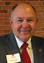 A smiling man wearing a suit and red tie stands in front of a brick wall. He has a nametag on his jacket.