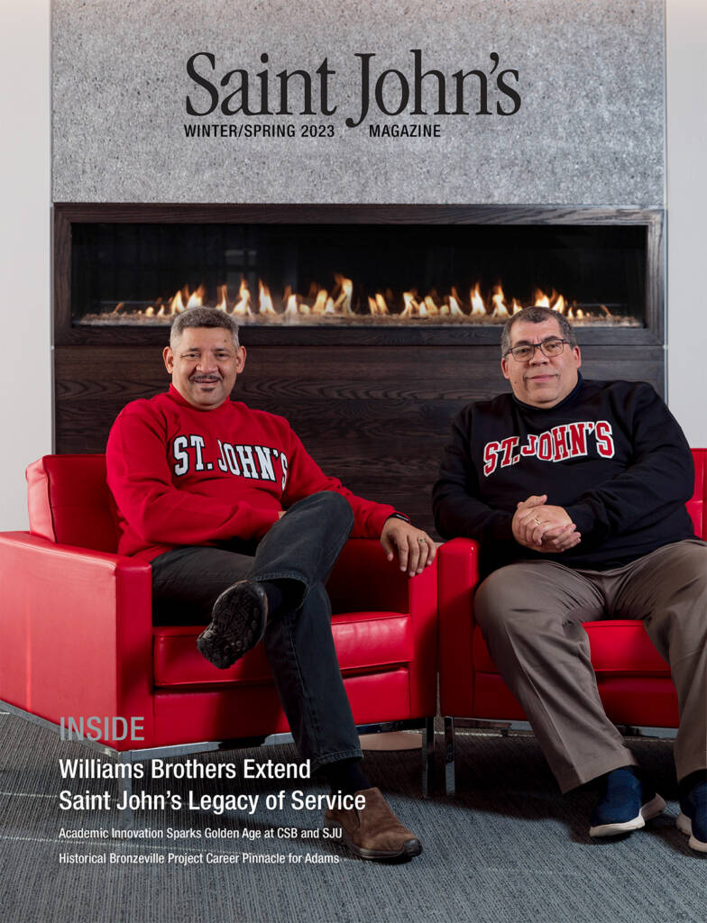 Two smiling men sitting in red armchairs, wearing red and blue "St. John's" sweatshirts, next to a modern fireplace. They are featured on the cover of Saint John's Magazine, Winter/Spring 2023 edition. Text highlights the Williams brothers' story.