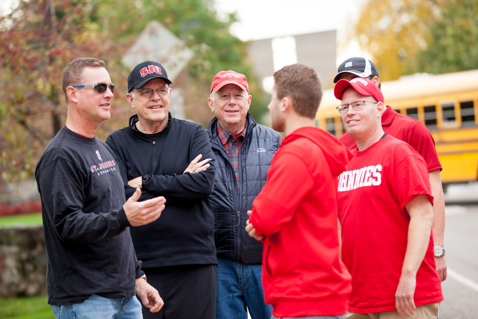 Five men in casual attire stand outdoors, talking and smiling. They wear red and black shirts, caps, and jackets. Two are holding crossed arms. A yellow school bus is visible in the blurred background.