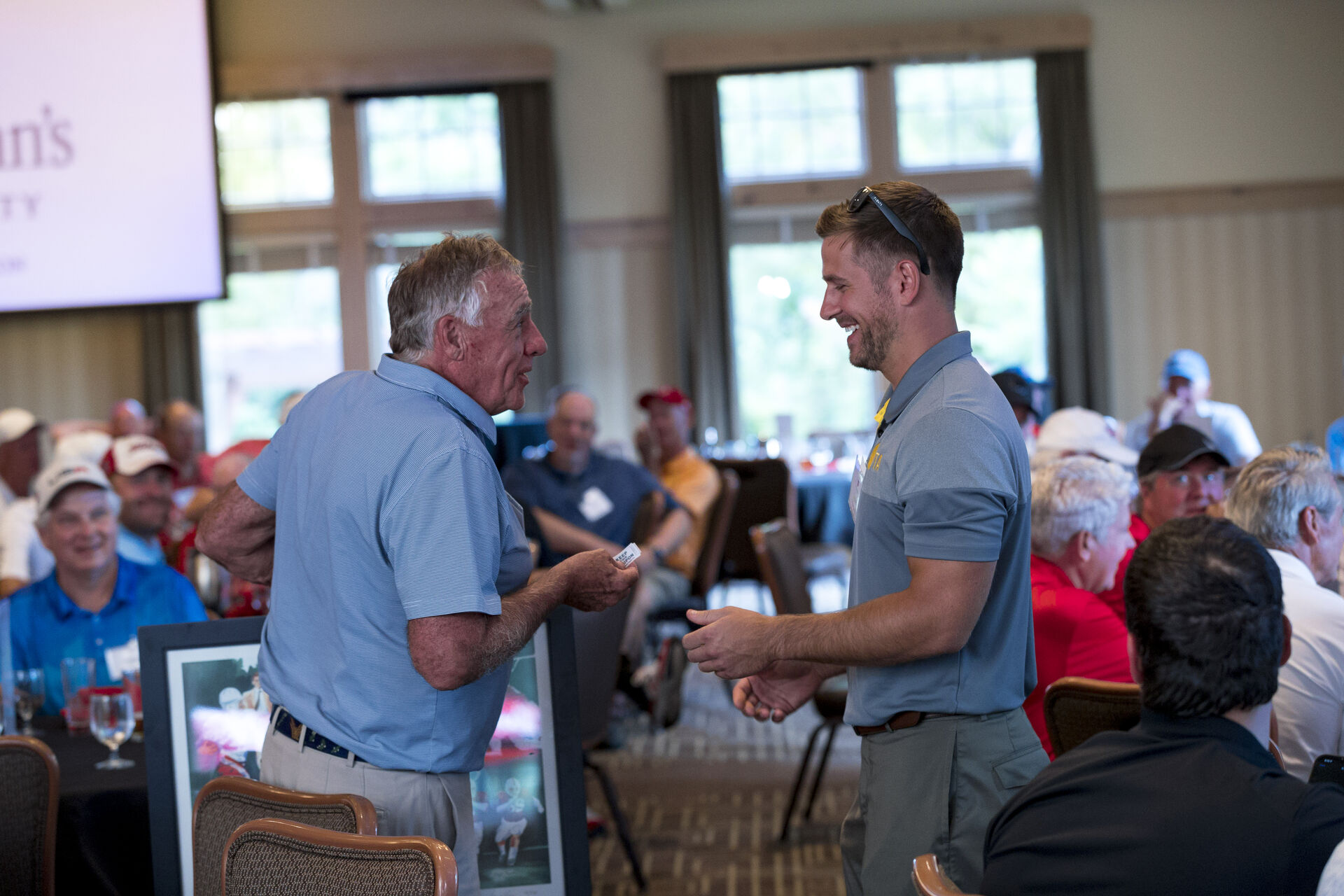 Two men stand and converse at a golf event in a room filled with seated attendees. The older man, holding papers, engages with the younger man, who is smiling. People in the background wear casual attire and hats, sitting around tables.