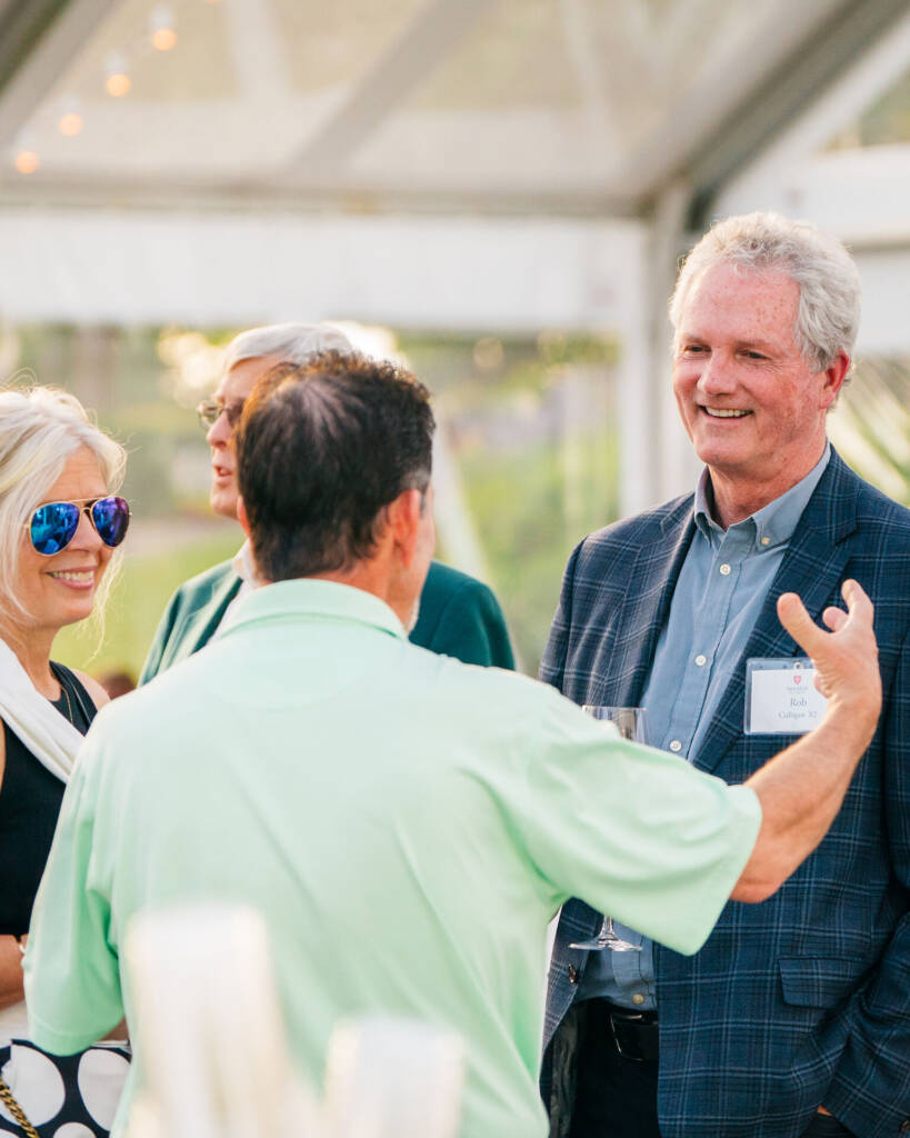 A group of people stands outside under a tent, engaging in conversation. An older man smiles, wearing a dark blazer with a name tag. A woman with long blonde hair, sunglasses, and a black-and-white outfit listens attentively. Warm, sunny ambiance.