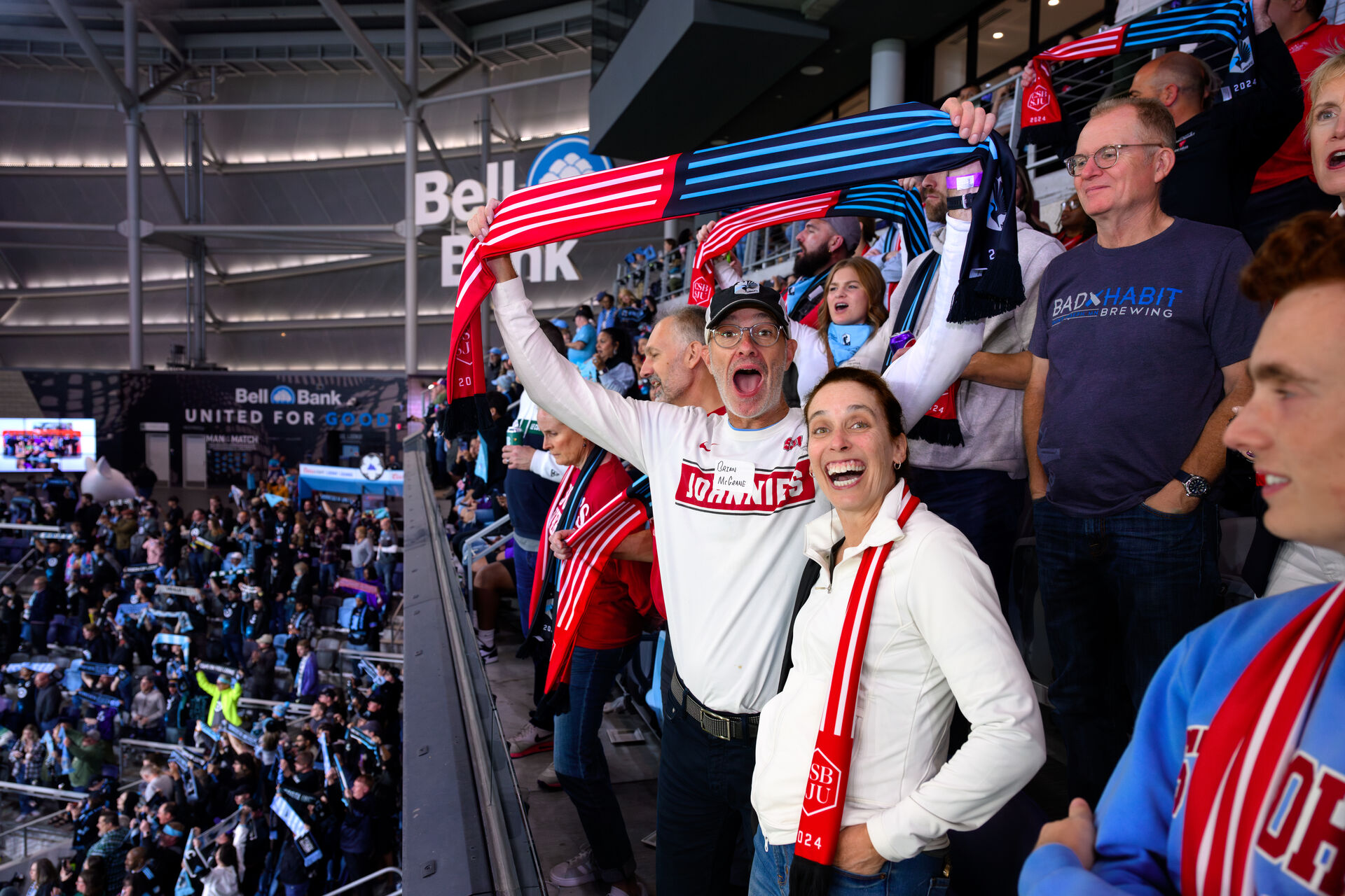 Fans in a stadium cheer enthusiastically, wearing scarves and shirts with team logos. One person in the foreground looks excited, surrounded by others who are standing and clapping. The stands are filled with more fans.