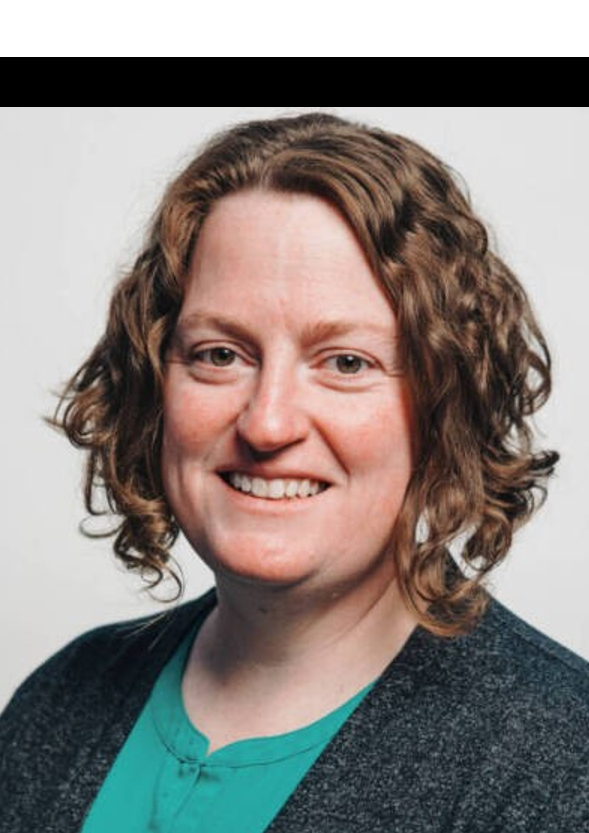 A person with shoulder-length curly hair, wearing a green shirt and a dark sweater, smiles while looking at the camera. The background is plain and light-colored.