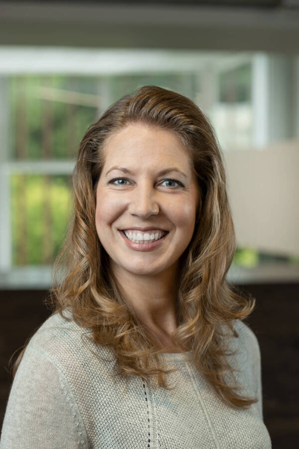 A woman with long wavy hair smiling at the camera. She is wearing a light gray sweater. The background is blurred, featuring a window and greenery.