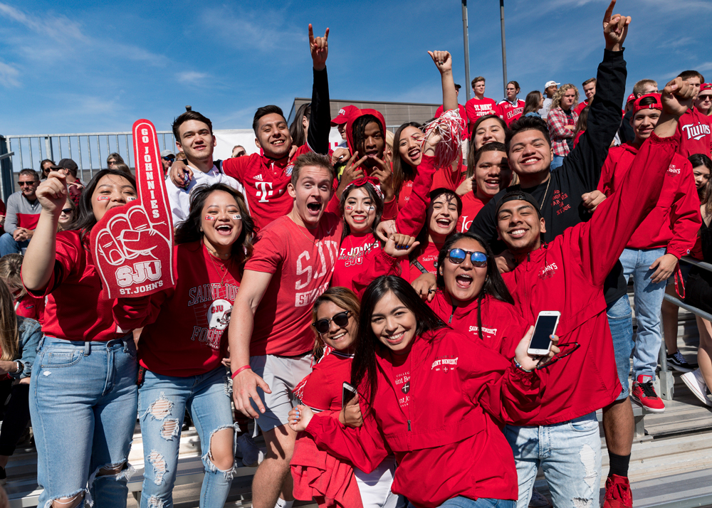 A large group of enthusiastic fans, mostly wearing red clothing with logos and text, smile and cheer in an outdoor stadium. One person holds a large red foam finger. The sky is clear, and the crowd is packed closely on bleachers, showing school spirit and excitement.