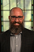 A man with a thick beard, wearing glasses, a gray shirt, and a dark blazer, stands in front of a window with a colorful stained glass design. He is smiling at the camera. The background is softly lit, emphasizing the window's pattern.