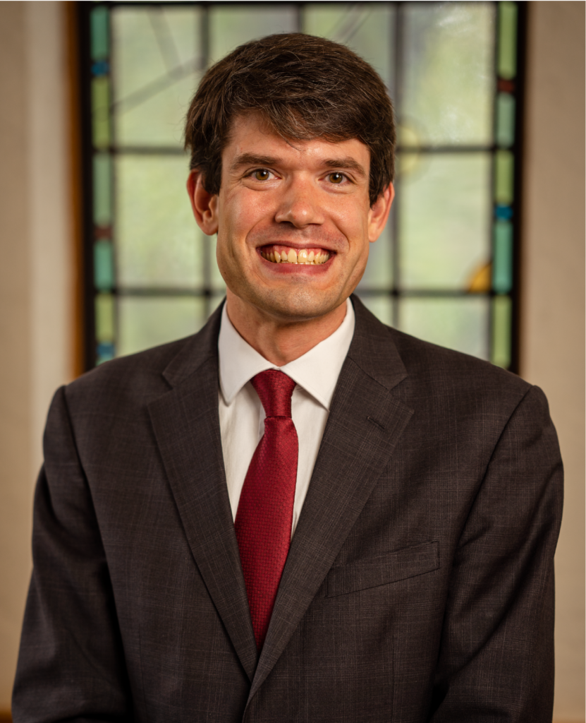 A man in a dark gray suit, white shirt, and red tie is smiling broadly while posing in front of a stained glass window. The background is softly blurred, emphasizing the vibrant colors of the stained glass.