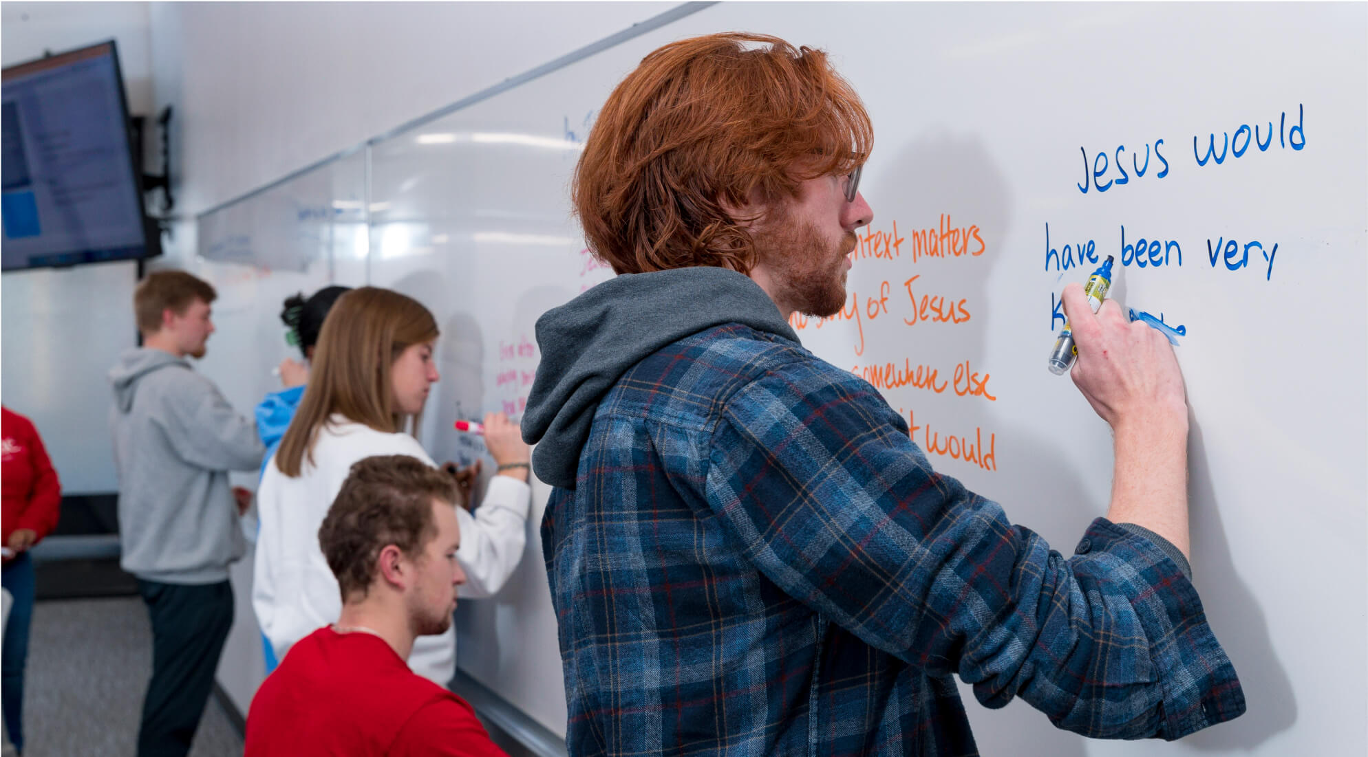 A group of students stands and sits near a whiteboard, writing and discussing. A student with red hair and wearing a plaid shirt focuses on writing in blue marker on the board. Others engage in conversation and writing, with classroom peripherals visible in the background.