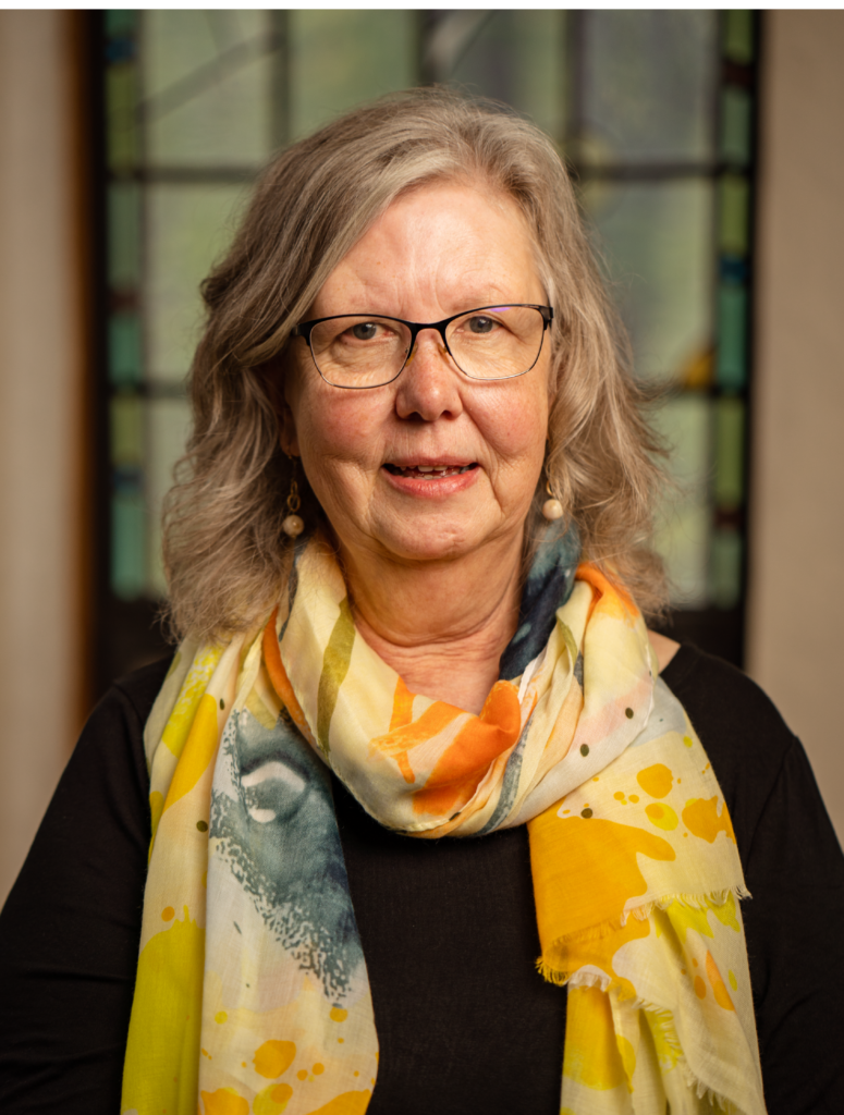 A woman with shoulder-length gray hair and glasses wearing a colorful scarf and black top is standing in front of a blurred background with a stained glass window. She is looking directly at the camera and smiling softly.