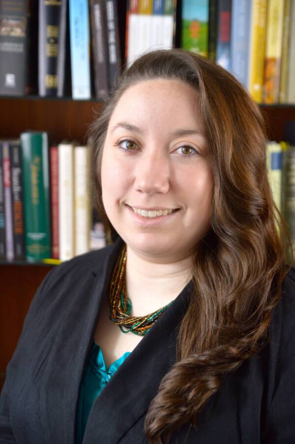 A person with long brown hair, wearing a dark blazer over a blue top, is smiling in front of a bookshelf filled with books.