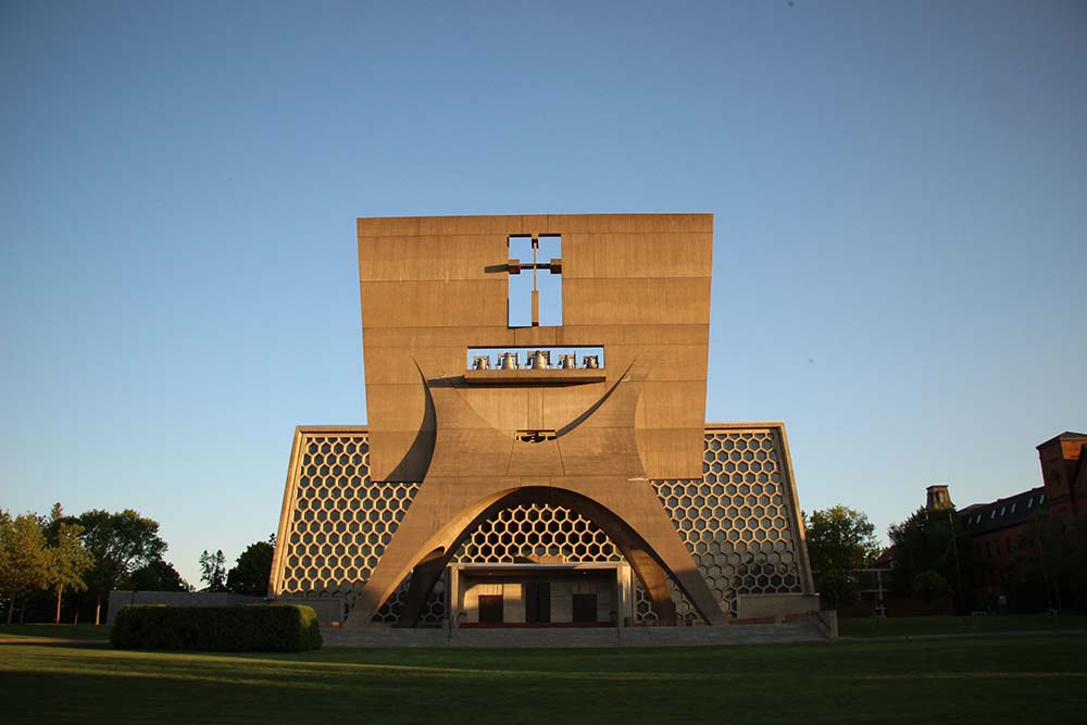 A concrete church with a unique modern design featuring large geometrical patterns and a prominent cross at the top of its façade. The sun is setting, casting a warm light on the building. Trees and a lawn surround the church, creating a serene atmosphere.