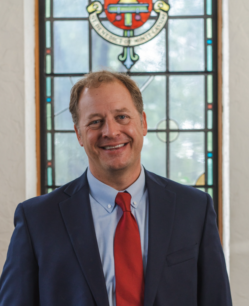 A man wearing a dark blue suit, light blue shirt, and red tie stands smiling in front of a multi-colored stained glass window. The window features a decorative design and symbols at the top. The room has light-colored walls.