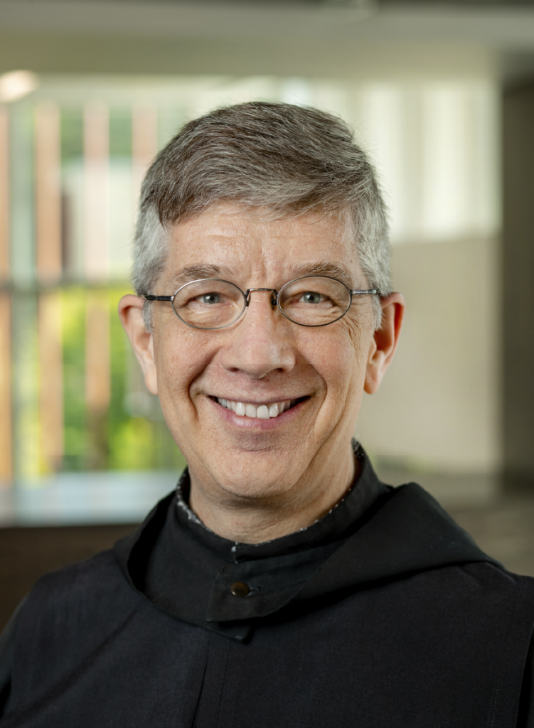 A smiling person with short gray hair and glasses, wearing a dark, formal robe. The background is softly focused, featuring a large window with natural light and some greenery visible outside.