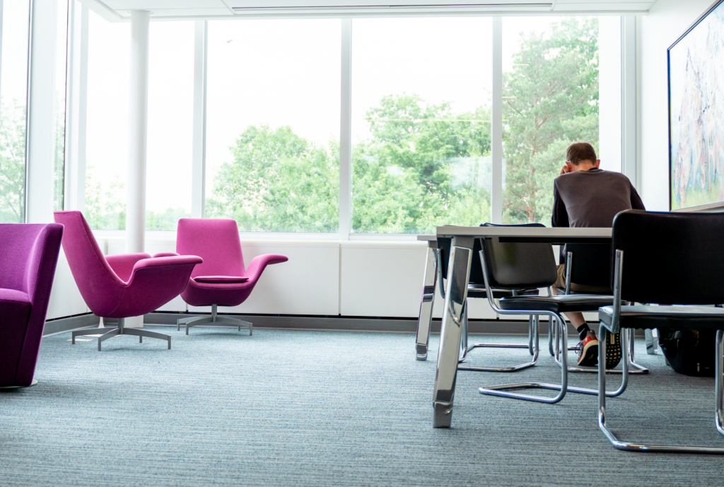 A person sits alone at a table in a modern, bright room with large windows. Two purple lounge chairs are positioned to the left, and black chairs are around the table. Outside, green trees are visible through the windows. The setting appears quiet and contemplative.