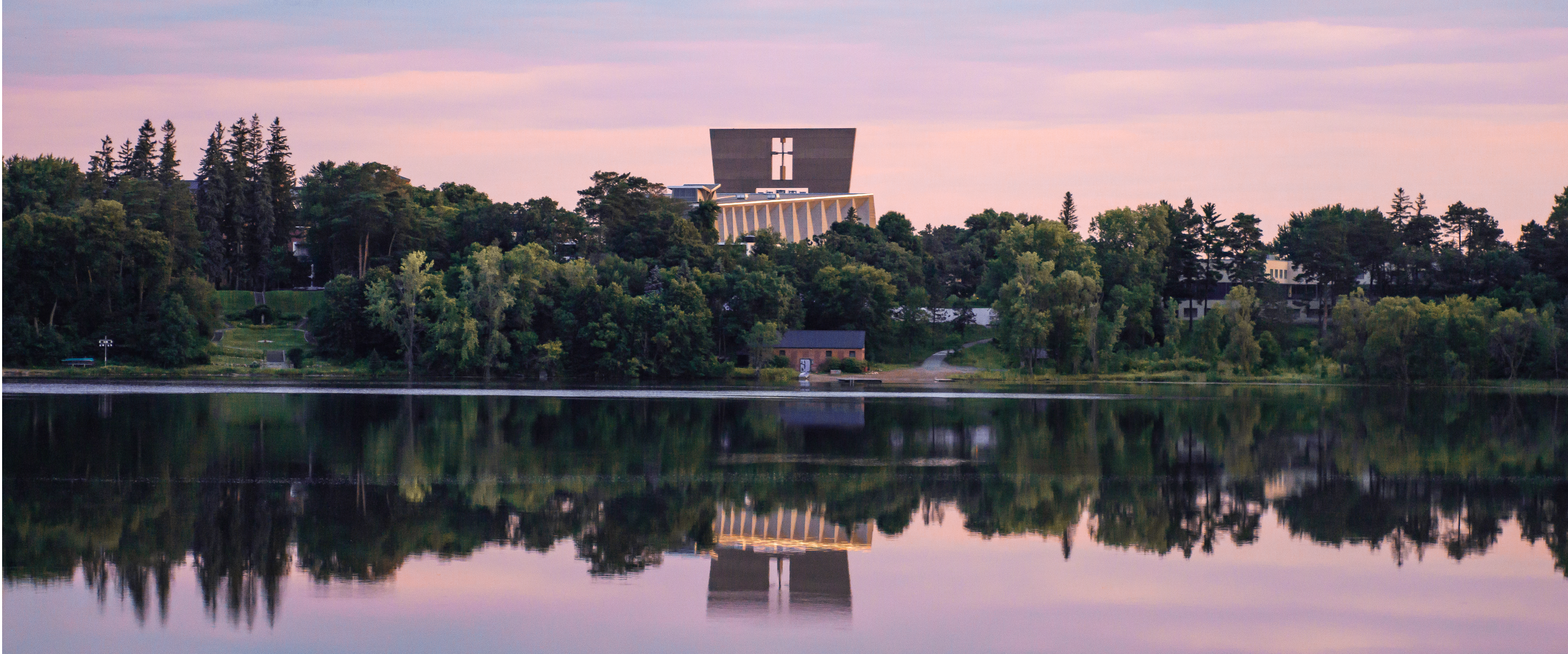 A serene lake reflects a forested shoreline and a large modern building with a distinct design, captured during the evening with a pinkish sky. Trees and greenery surround the water and shoreline, creating a tranquil and picturesque scene.