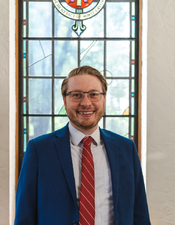 A person wearing glasses, a blue suit, and a red-striped tie stands smiling in front of a stained glass window. The background is slightly out of focus, highlighting the intricate window details.