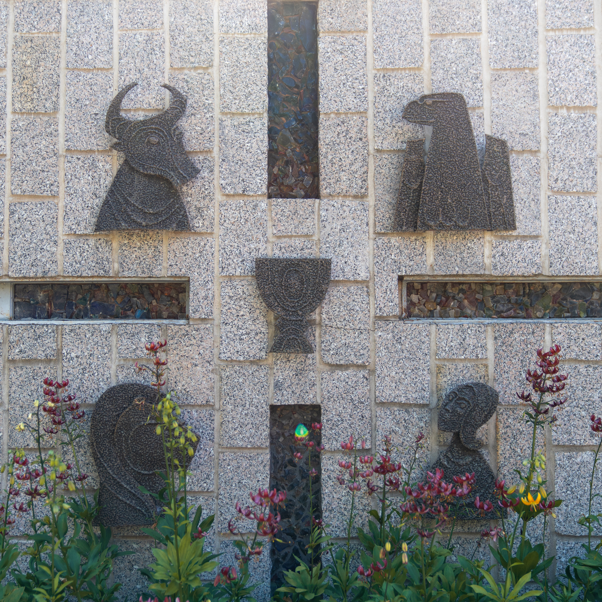 A wall features four ornate stone carvings: a bull, an eagle, a chalice, and a lion. Each carving is evenly spaced and set against a textured backdrop with vertical glass insets. In front, colorful flowers add to the scene's vividness.