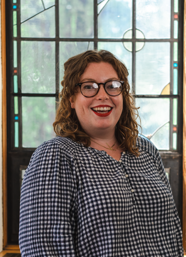 A smiling person with curly, shoulder-length hair and glasses stands in front of a window with a stained glass pattern. They are wearing a black and white checkered blouse.