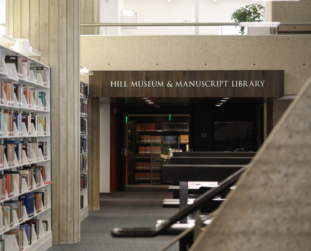 The image shows the interior of a library with several bookshelves filled with books on the left side. A sign above an entrance reads "Hill Museum & Manuscript Library." There is a partial view of a staircase and tables in the foreground.