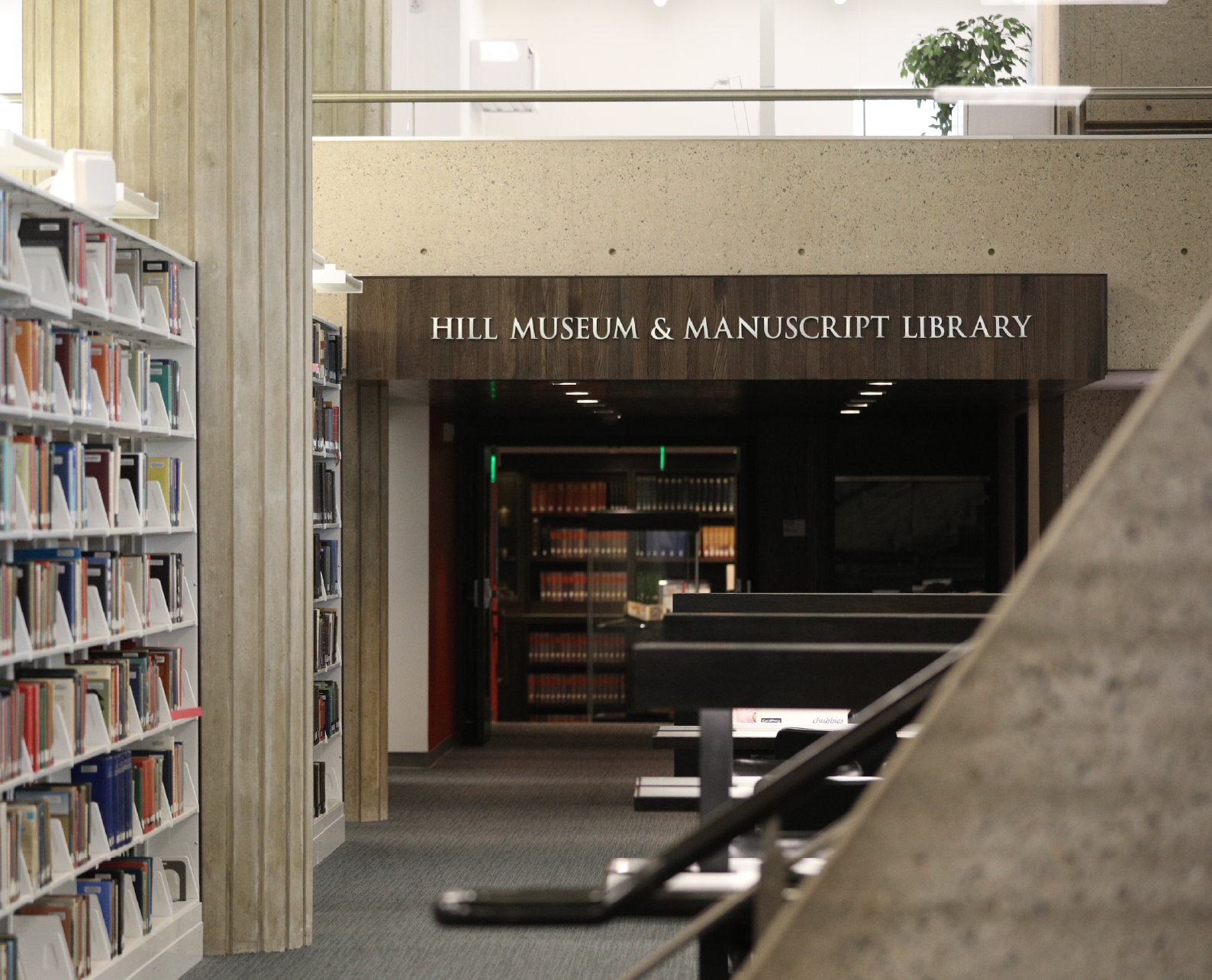 The image shows the interior of a library with several bookshelves filled with books on the left side. A sign above an entrance reads "Hill Museum & Manuscript Library." There is a partial view of a staircase and tables in the foreground.