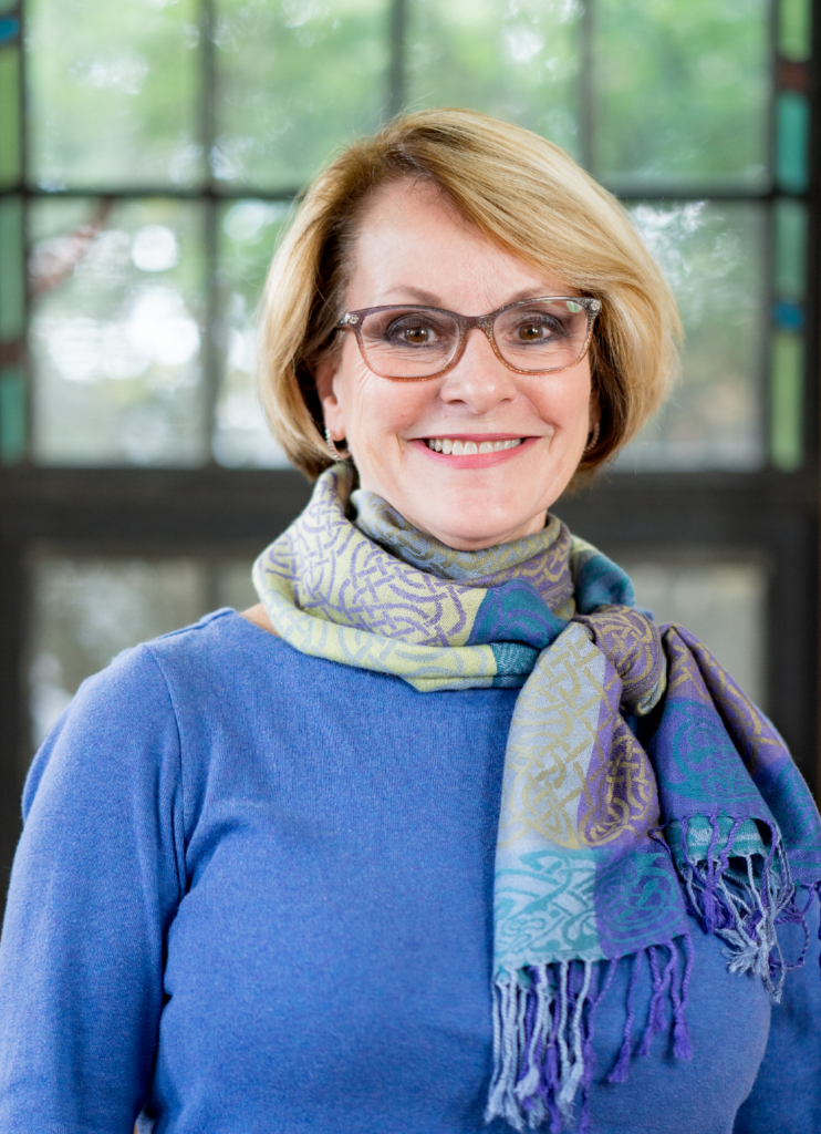 A smiling woman with short blonde hair, wearing glasses, a blue sweater, and a multicolored scarf stands in front of a window with a blurry outdoor view.